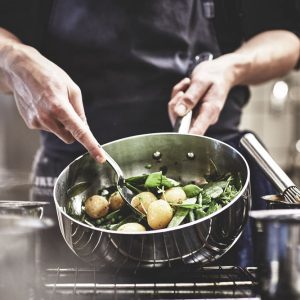 Händer som rör om potatis och grönsaker i en stekpanna.